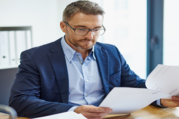 Image of a business man seated at a desk. He is wearing glasses and reviewing a lengthy document, which is secured by a paperclip