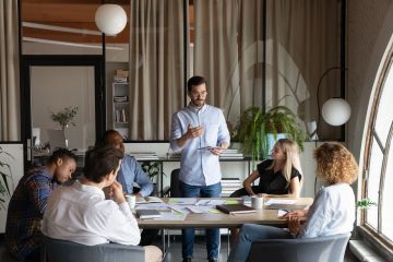 Meeting - man leading a team meeting with six people seated around a table in a modern office.