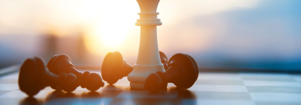 A white chess king on a chessboard, surrounded by fallen black pawns. The setting sun in back is casting a warm glow.